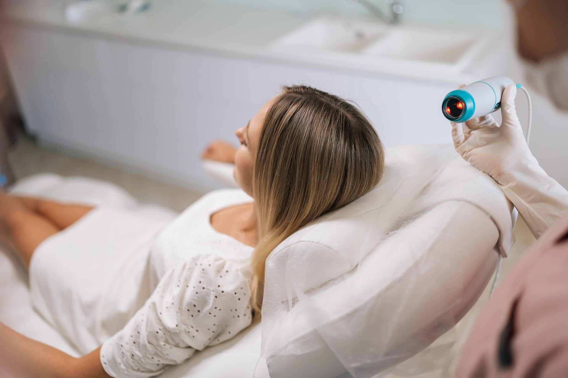 Unrecognizable trichologist in gloves and mask performing hair treatment procedure using spectrum phototherapy device. Blonde female patient having hair light therapy